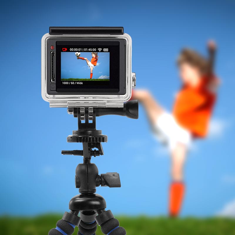 A Mini Tripod Mount for GoPro HERO Action Cameras keeps the camera steady as it records a person in an orange shirt kicking a soccer ball. The subject is blurred in the background but sharp on the cameras screen against a bright blue sky.