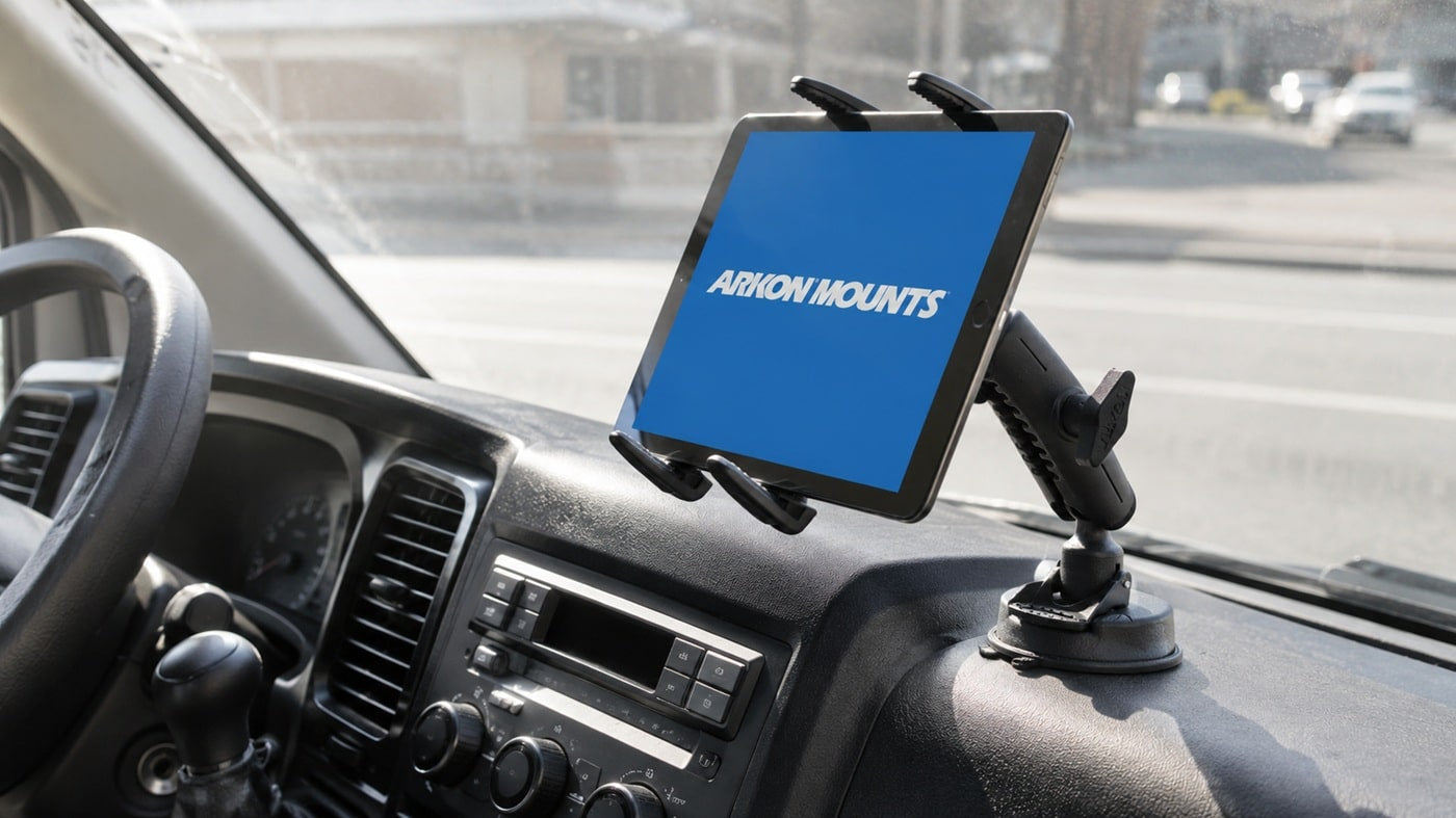 Image of a tablet holder mounted to a dashboard with the ARKON Mounts logo on the screen in white writing and blue background. In the backgorund, ther is a road, cars and buildings.