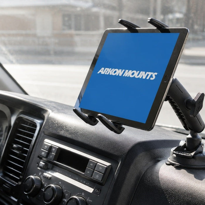 Image of a tablet holder mounted to a dashboard with the ARKON Mounts logo on the screen in white writing and blue background. In the backgorund, ther is a road, cars and buildings.
