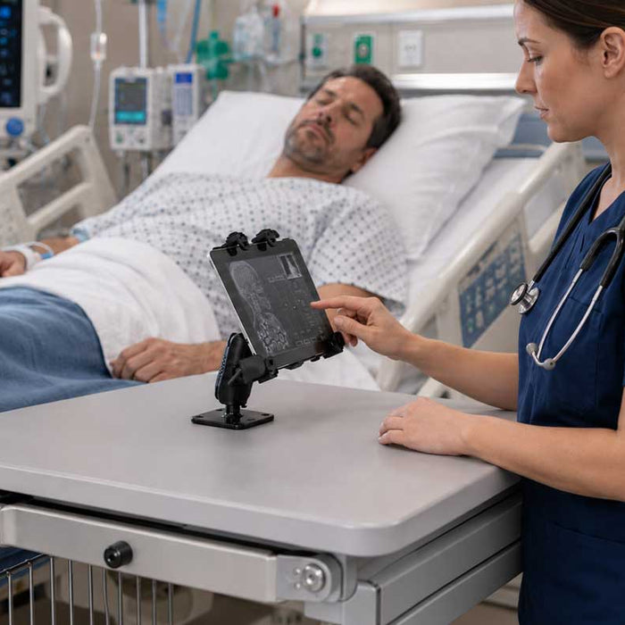 Image of a white female nurse in dark blue scrubs working on a mounted tablet that's attached to a healthcare cart. She is in front of a hospital bed with a while male with dark hair. There is a screen showing his vitals in the background. 
