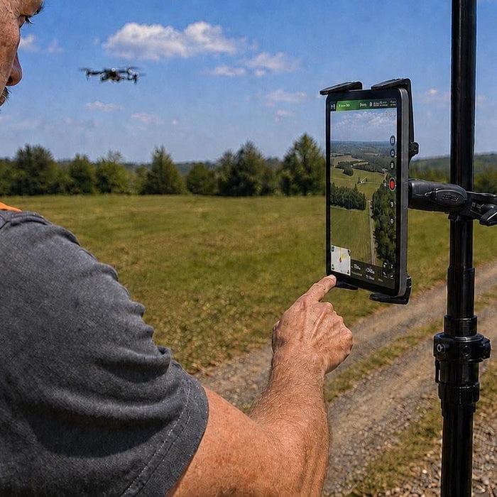 Image of a man working on a tablet that is attached to a pole. He is on a track with grass in the background and a drone is flying in the sky