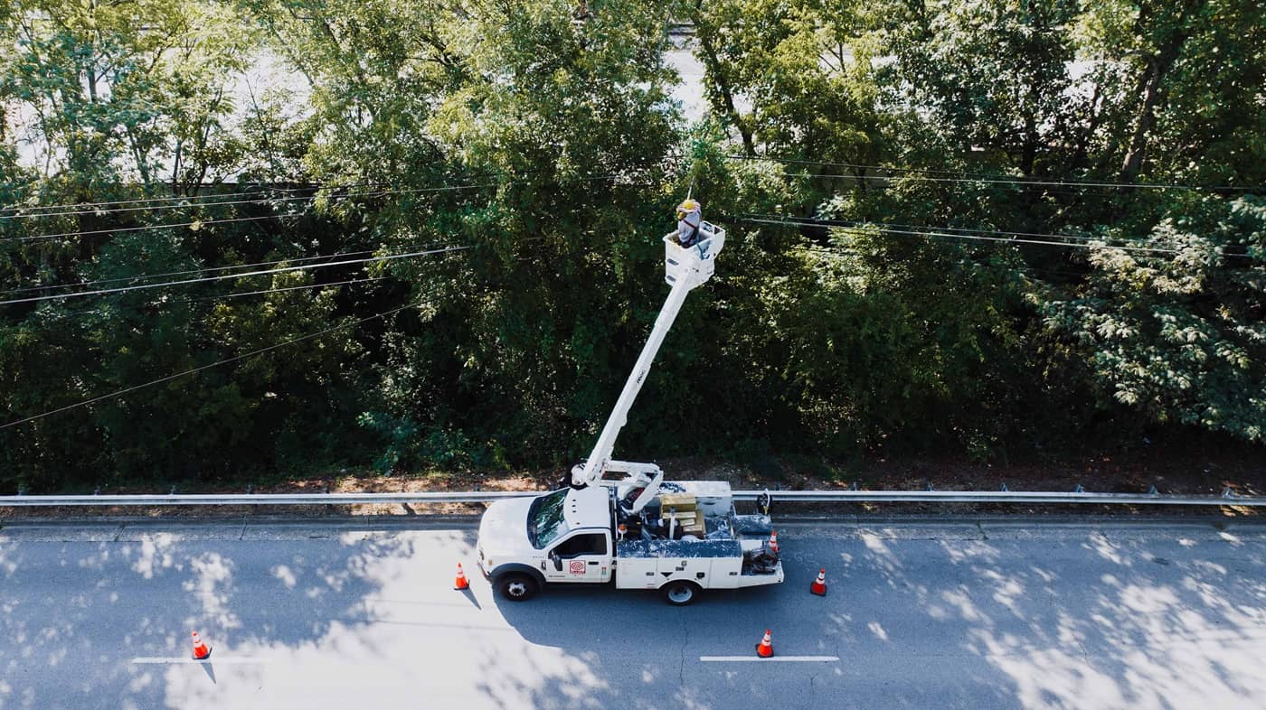 Image of a white service truck with cherry picker parked on a road. The service technician is working on some overhead cables. In the background there are a row of large tress. The angle of the image is looking down at an angle