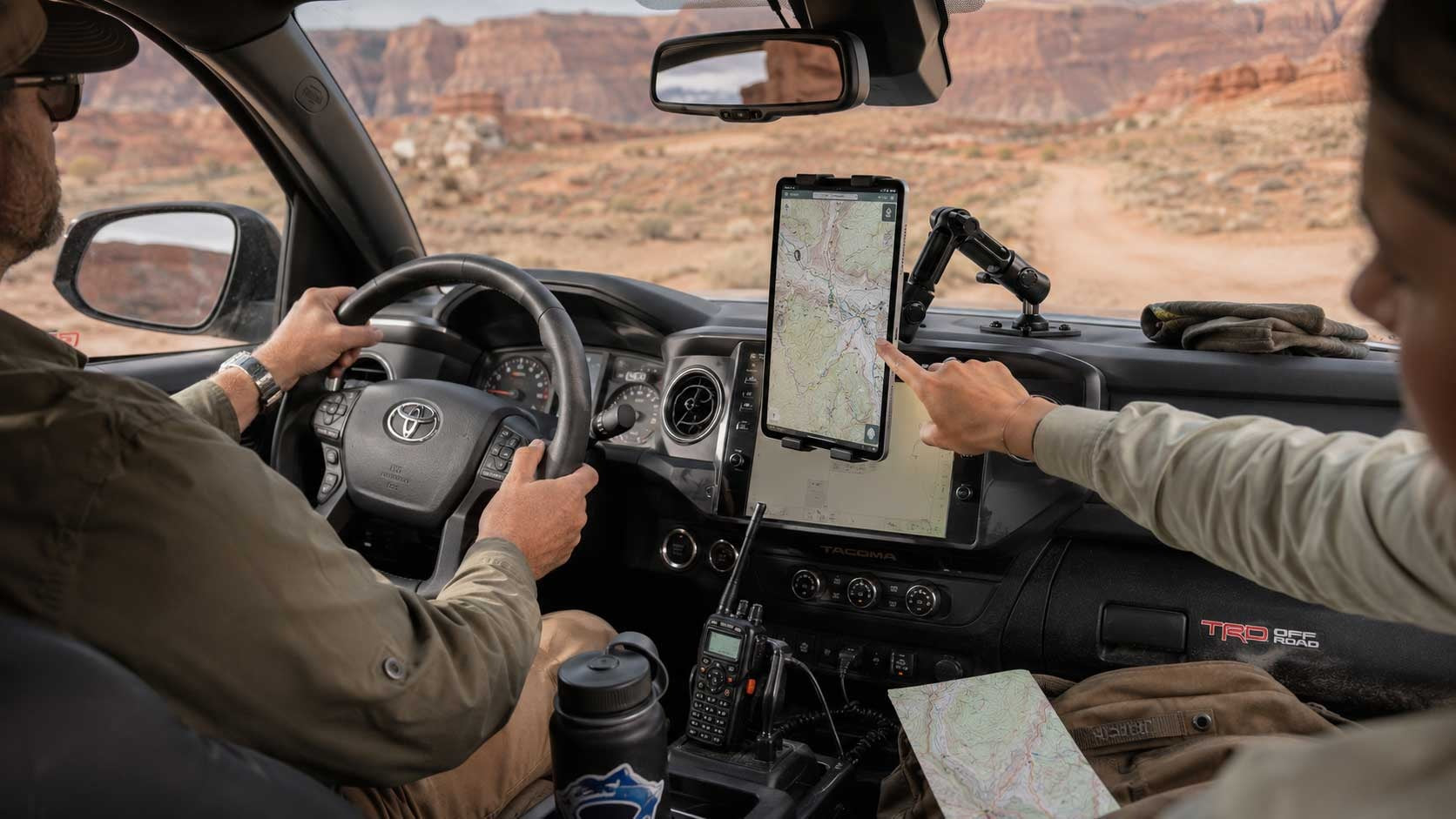Image of inside a truck with a man driving and someone else pointing to a mounted tablet on the dashboard. There is a desert and roacky mountains in the background.