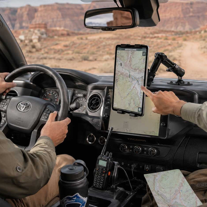 Image of inside a truck with a man driving and someone else pointing to a mounted tablet on the dashboard. There is a desert and roacky mountains in the background.