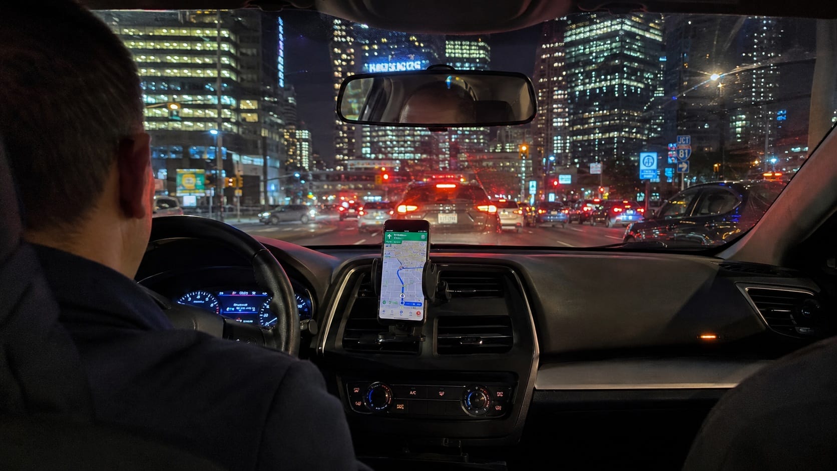 Image of the inside of a rideshare showing the back of the driver's head and shoulders. There is a mounted Smartphone on the console showing GPS. Out the windshield is other cars and skyscrapers. It is night time.
