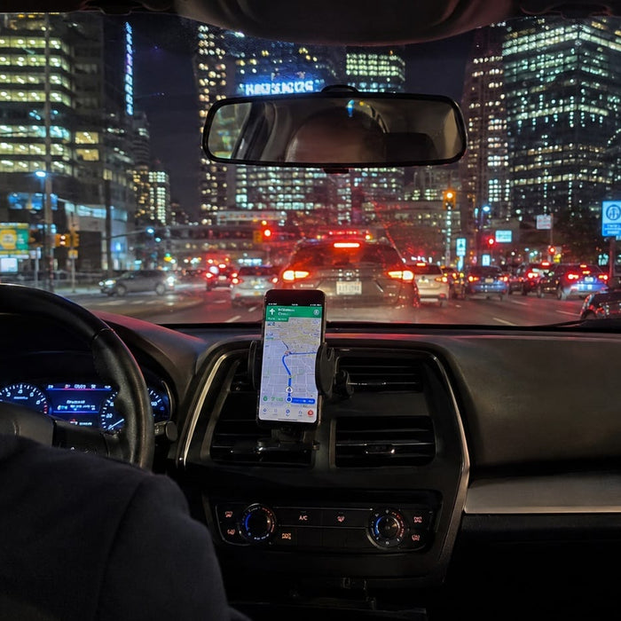 Image of the inside of a rideshare showing the back of the driver's head and shoulders. There is a mounted Smartphone on the console showing GPS. Out the windshield is other cars and skyscrapers. It is night time.