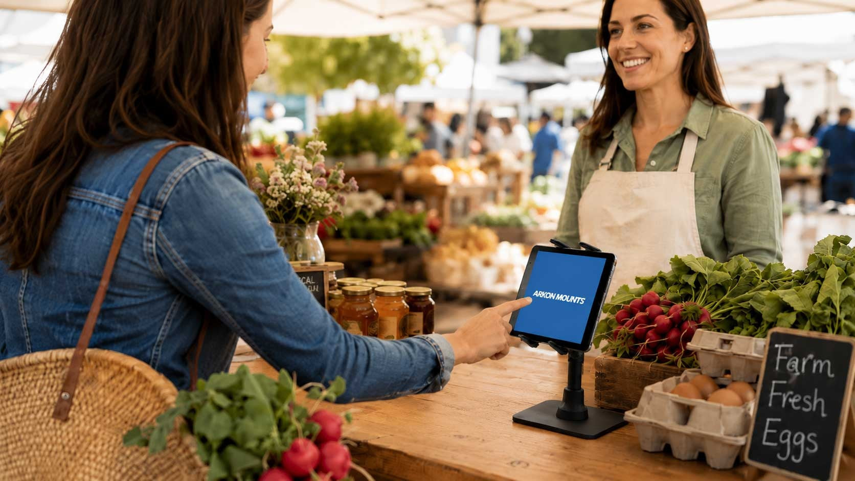 Image of two women at a farmers' market. One woman is pressing on a tablet that's on a tablet stand on the countertop. There are eggs and fress produce also on the countertop.