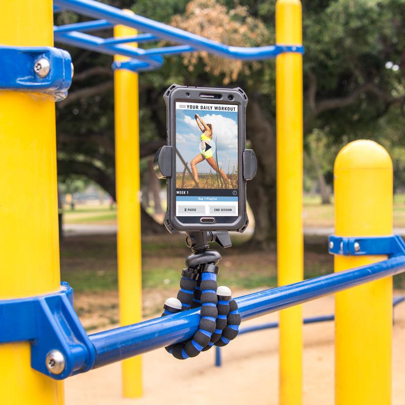 The 11 inch Tripod with RoadVise® Phone Holder Mount for Video is used outdoors on blue playground bars to hold a smartphone displaying a workout video, with trees and greenery in the background.