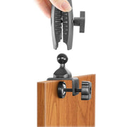 A hand attaches the 25mm Swivel Ball to C-Clamp Mounting Pedestal to the edge of a wooden desk using its screw-tightened clamp.