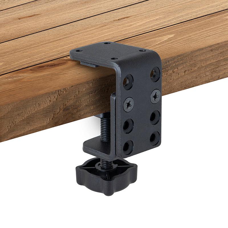 A black 4-Hole AMPS to Adjustable Clamp Adapter is attached to the edge of a wooden surface, secured with screws and a large plastic tightening knob underneath.