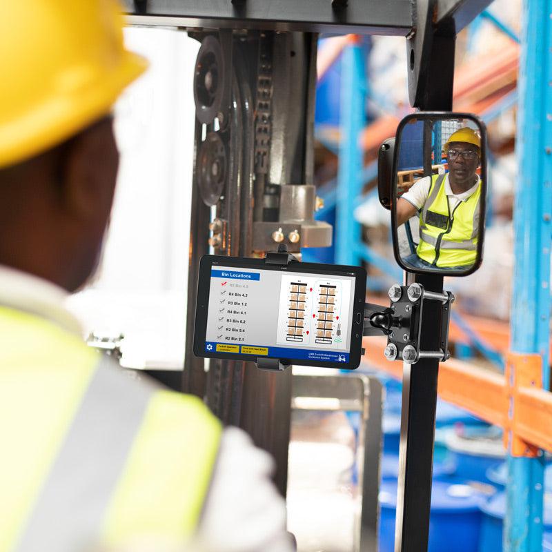 A worker in a yellow hard hat and vest operates a forklift in a warehouse, viewing inventory data on his screen secured by the RoadVise® Ultra Forklift Front Guard Phone and Tablet Mount, with his image reflected in the rearview mirror.
