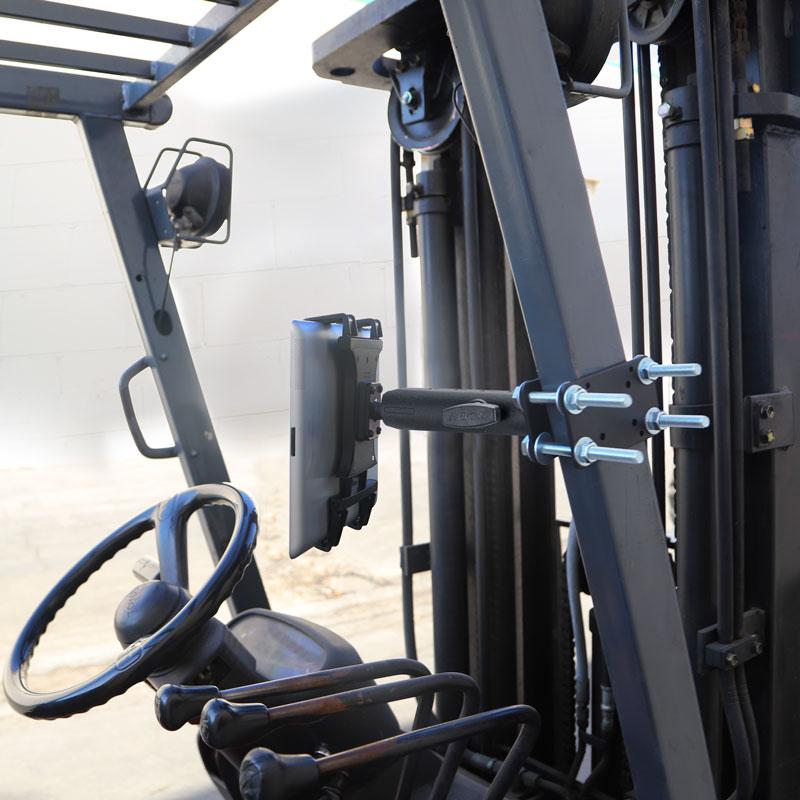 Close-up of a forklift cockpit featuring the 7.25 inch Metal Robust Forklift Front Guard Slim-Grip® Tablet Mount, securely holding a tablet next to the steering wheel and levers. A white brick wall is visible in the background.