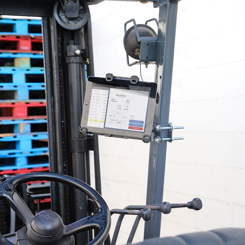 The LockVise™ Locking Forklift Front Guard Tablet Mount with 7.25 Shaft displays data on a forklift parked near red and blue pallets against a white wall, with the steering wheel and control levers visible in the foreground.