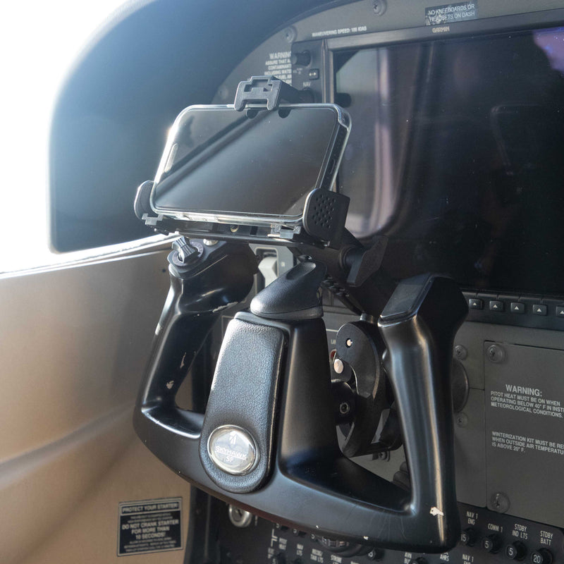 A SkyHold™ Yoke Locking Phone Mount secures a smartphone to an aircraft control yoke, positioned in front of flight instruments and displays, with cockpit controls and warning labels visible in the background.