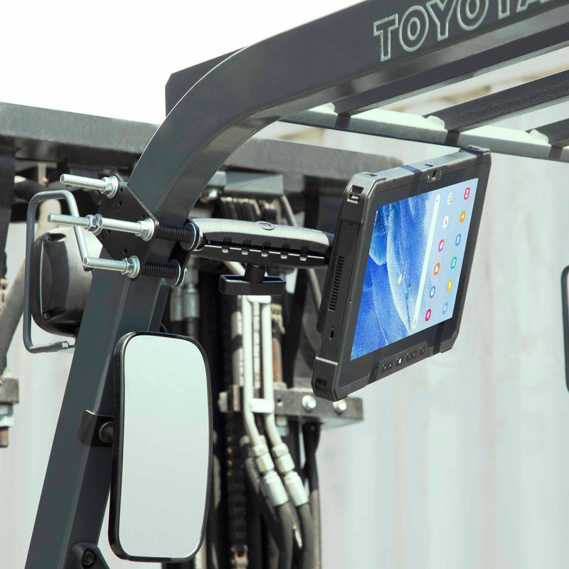 A 10.25 inch Metal Robust Forklift Front Guard VESA Mount secures a rugged tablet to the forklift’s metal frame near the rearview mirror, displaying app icons on a blue screen, with hydraulic hoses and outdoor light visible in the background.