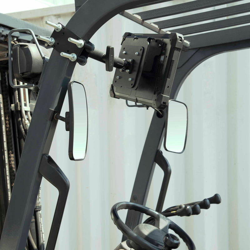 A close-up of a forklift cabin displays the steering wheel, control levers, side mirrors, and a 10.25 inch Metal Robust Forklift Front Guard VESA Mount attached to the overhead guard, with a corrugated metal wall in the background.