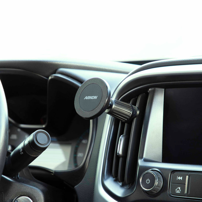 A Magnetic Phone Holder with Vent Mount - MagSafe Compatible is secured to a car’s air vent near the dashboard, with part of the steering wheel and infotainment screen visible.
