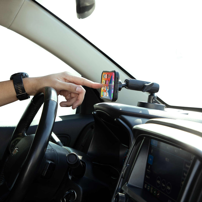 A person touches a smartphone on the Magnetic Phone Holder with Suction Mount - MagSafe Compatible, showing its home screen; the steering wheel and infotainment system are also visible.