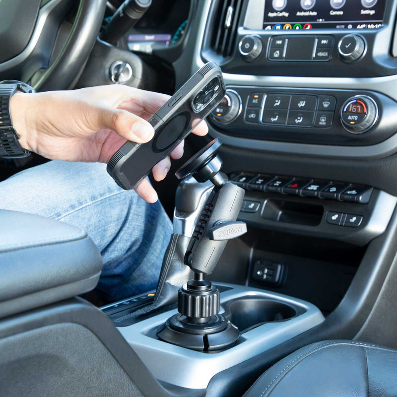 A person holds a smartphone near the Magnetic Phone Holder with Car Cup Mount - MagSafe Compatible, attached beside a car’s gear shift inside a modern vehicle, highlighting the dashboard and controls.