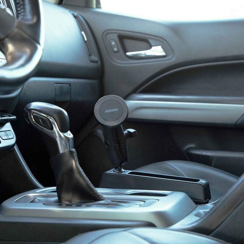 A close-up of a car’s interior shows the Magnetic Phone Holder Console Wedge Mount – MagSafe Compatible attached to the center console near the front passenger seat, with the gear shift, dashboard, and steering wheel partially visible.