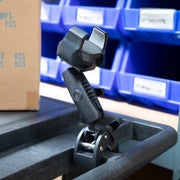 The RoadVise® Ultra Holder with Clamp Mount, in black, is attached to a carts edge. Blue plastic storage bins and a cardboard box are visible in the background.
