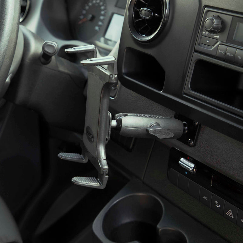 A close-up of a car interior shows the Slim-Grip® Tablet Holder with Metal Drill Base Mount installed on the dashboard near the steering wheel and air vent, with buttons and cupholders visible.
