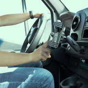 A person sitting in a car adjusts the Slim-Grip® Tablet Holder with Metal Drill Base Mount near the dashboard while holding the steering wheel with their left hand.