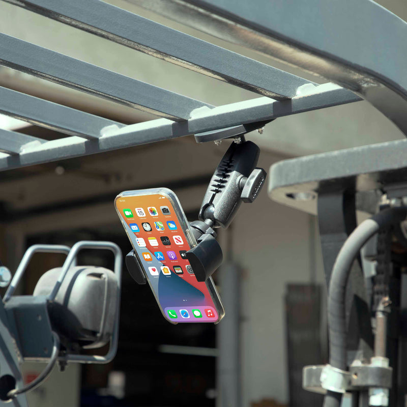A RoadVise® XL Phone and Midsize Tablet Magnetic Mount holds a smartphone upside down on a metal bar near industrial equipment outdoors, with the phone displaying app icons.