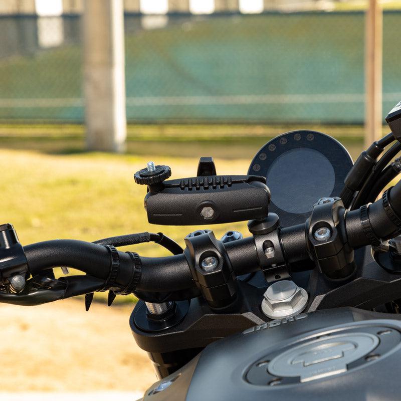 Close-up of a motorcycle handlebar featuring the Aluminum Motorcycle Camera Handlebar Mount with 2.75 Shaft, alongside control buttons and part of the digital display, set against a blurred fence and grassy background.