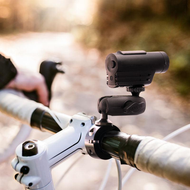 An Aluminum Motorcycle Camera Handlebar Mount with 2.75 Shaft holds a black action camera on a white bike, as a persons hand grips the handlebar outdoors in warm sunlight.