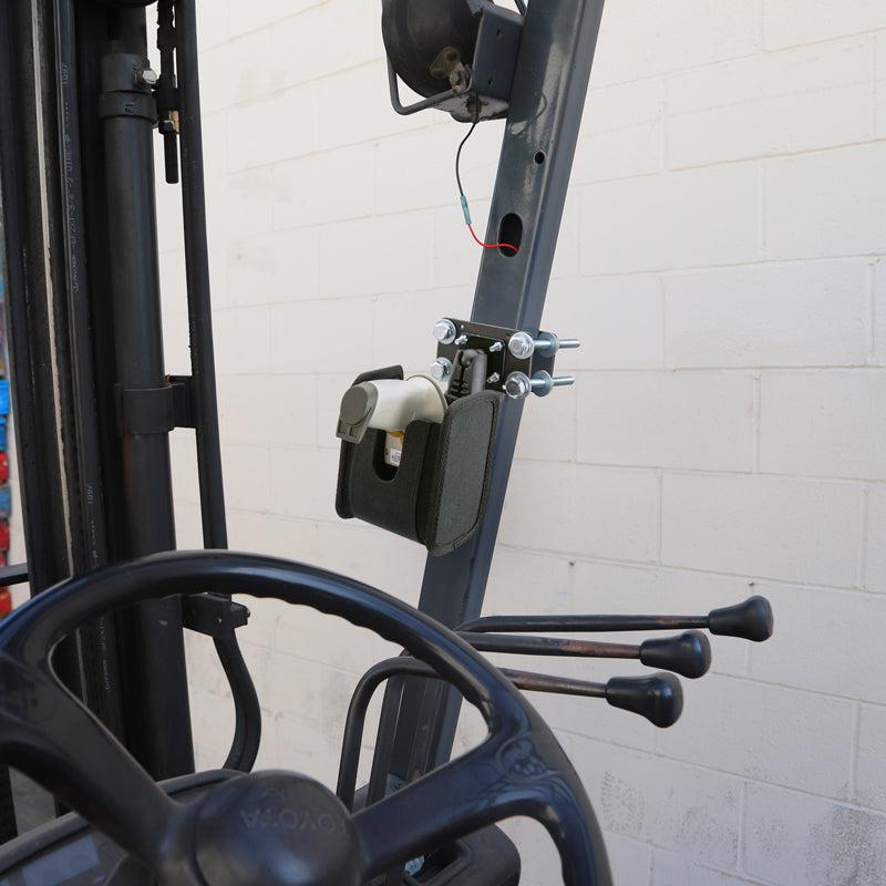 The Barcode Scanner Universal Forklift Front Guard Mount holds a barcode scanner and hand sanitizer dispenser beside the steering wheel and levers of a forklift, set against a white brick wall for efficient warehouse fulfillment.