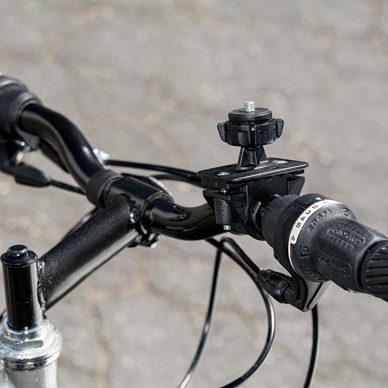 Close-up of a Bike or Motorcycle Handlebar Camera Mount for Canon, Sony, Samsung, Panasonic, Nikon Cameras with a 1/4-20 mounting pattern attached to a black bicycle handlebar. A gear shifter is on the right; blurred pavement is in the background.