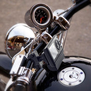 Close-up of a motorcycle handlebar with a speedometer, chrome finish, and a Bike or Motorcycle Handlebar Mount for XM and Sirius Satellite Radio (4-Hole AMPS pattern) securing a digital device. Building reflections appear in the metal surfaces.