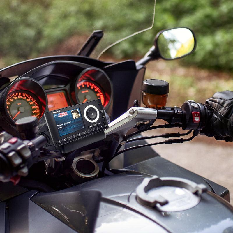 A close-up shows a motorcycle dashboard with the Bike or Motorcycle Handlebar Mount for XM and Sirius Satellite Radio using a 4-Hole AMPS pattern. Willie Nelson’s song plays on the radio; greenery blurs in the background as a gloved hand grips the right handlebar.
