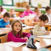 A young girl in a classroom smiles at the camera as she writes in her notebook. Her tablet is propped up on the desk using a Portable Tablet/Phone Stand. Other children are busy working in the background.