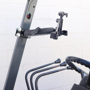 The LockVise™ Locking Forklift Front Guard Tablet Mount with a 10.25 inch Metal Robust™ Shaft is secured to a metal frame beside control levers on industrial equipment, with a white brick wall in the background.