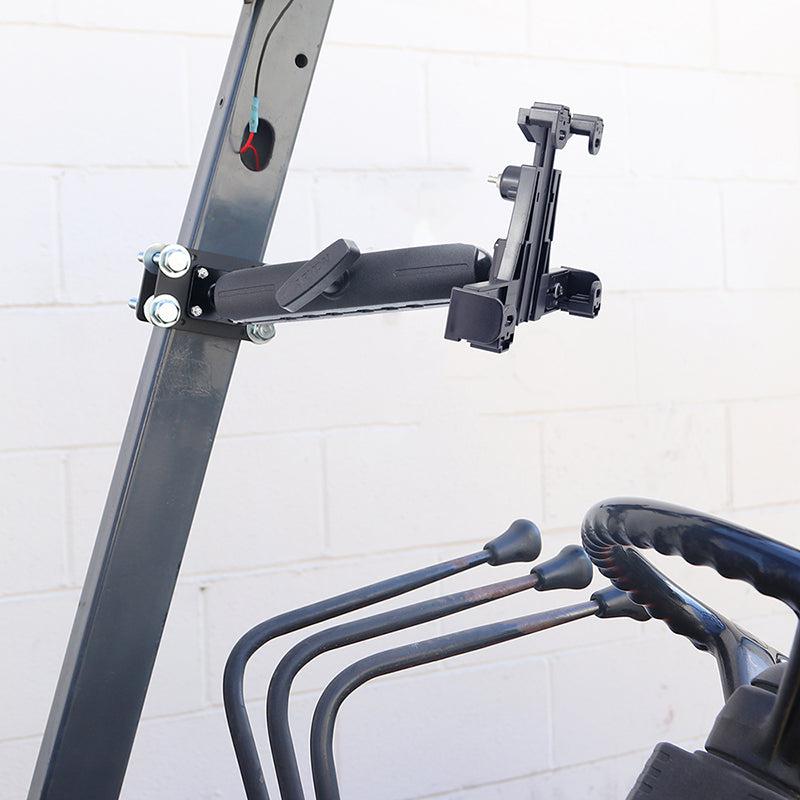 The LockVise™ Locking Forklift Front Guard Tablet Mount with a 10.25 inch Metal Robust™ Shaft is secured to a metal frame beside control levers on industrial equipment, with a white brick wall in the background.
