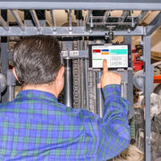 A man in a blue and green plaid shirt operates a forklift, using the LockVise™ Metal Locking Overhead Guard Tablet Mount with Multi-Pivoting Arm to secure his tablet, with warehouse items visible in the background.
