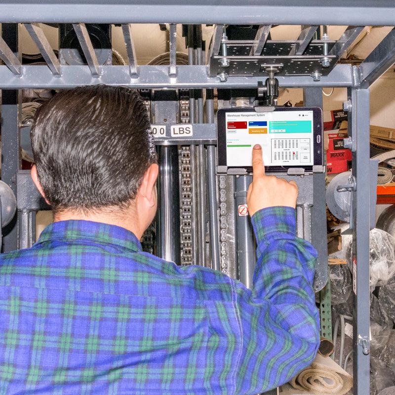 A man in a blue and green plaid shirt operates a forklift, using the LockVise™ Metal Locking Overhead Guard Tablet Mount with Multi-Pivoting Arm to secure his tablet, with warehouse items visible in the background.