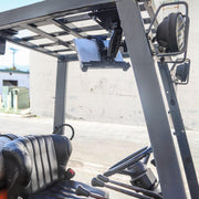 A close-up of a forklift cabin shows the driver’s seat, steering wheel, and a LockVise™ Locking Forklift Overhead Guard Tablet Mount with Robust™ Adjustable Arm under the roof, plus a headlight visible and sunlight streaming in.