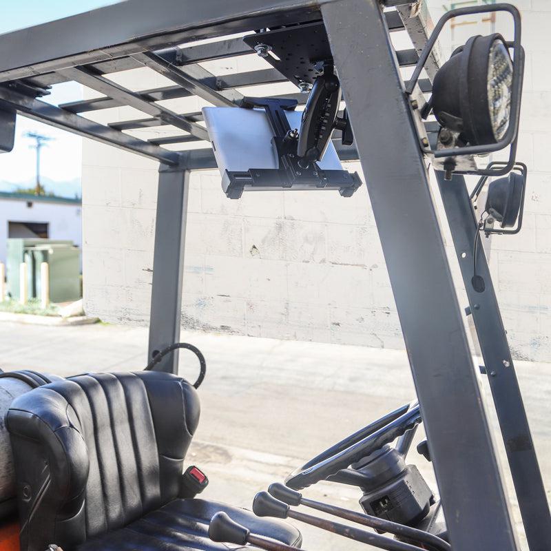 A close-up of a forklift cabin shows the driver’s seat, steering wheel, and a LockVise™ Locking Forklift Overhead Guard Tablet Mount with Robust™ Adjustable Arm under the roof, plus a headlight visible and sunlight streaming in.
