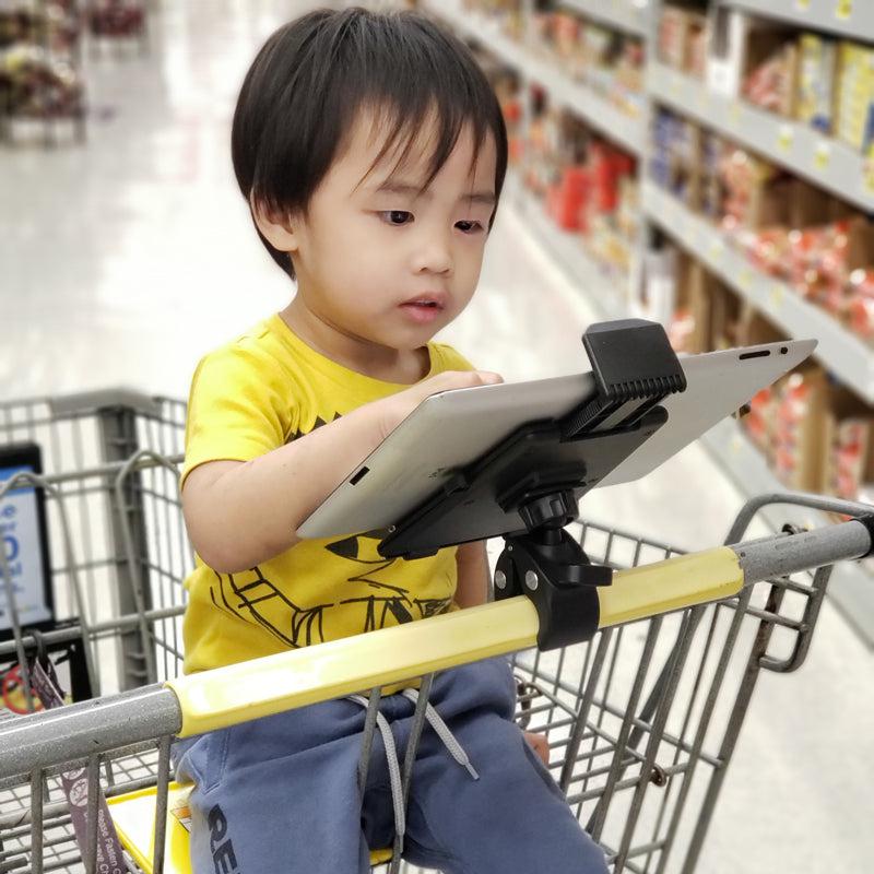 A young child in a yellow shirt and blue pants sits in a grocery cart, watching a tablet held by the Heavy-Duty Clamp Post Tablet Mount for iPad, Note, Tab and more attached to the handle. Store shelves are visible in the background.