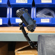 A Slim-Grip® Tablet Holder with Drill-Base Mount and 8 Multi-Angle Arm secures a rugged tablet at the edge of a workbench, with blue storage bins and cardboard boxes visible in the background.