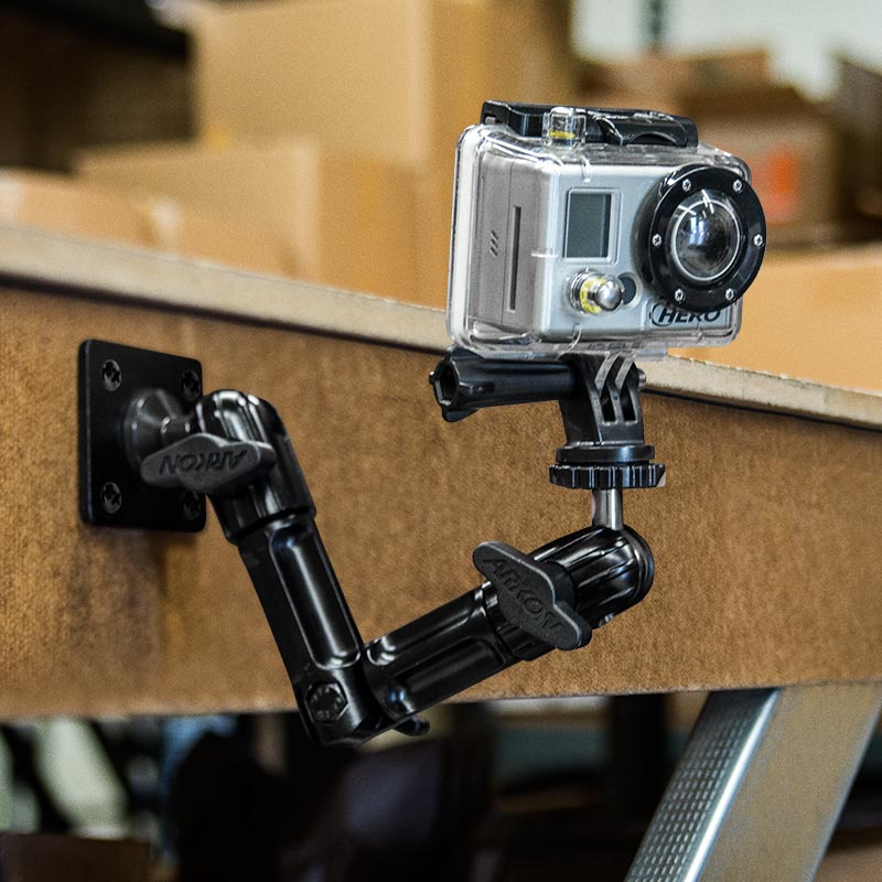 A GoPro HERO is attached to a table edge in a workshop or storage area using the Heavy-Duty Wall Mount with Multi-Angle Arm for GoPro Action Cameras, with cardboard boxes seen in the background.