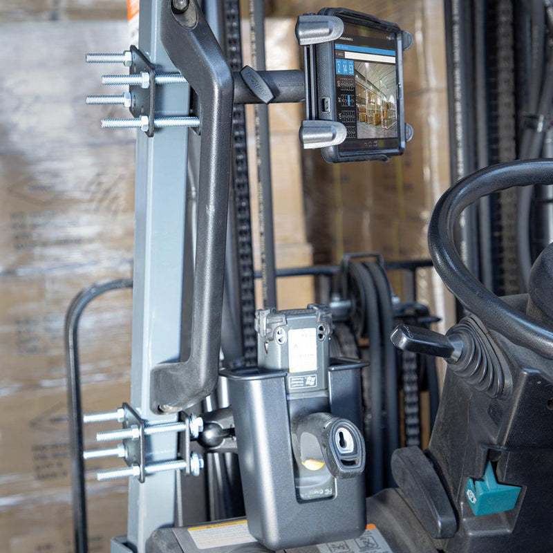 A close-up of a forklifts control area shows a Large Barcode Holster Forklift Front Guard Mount with 5 Shaft securing a digital device, with stacked cardboard boxes visible in the warehouse background.
