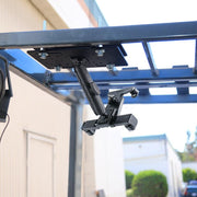 The LockVise™ Locking Forklift Overhead Guard Tablet Mount with 6 metal shaft is attached beneath a blue metal rack outdoors, with adjustable arms—perfect for securely holding tablets or electronic devices.
