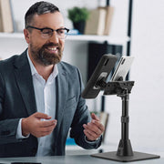 A bearded man in a suit and glasses sits at a desk, smiling and gesturing while using his smartphone on the Dual Phone Magnetic Mount Desk Stand in a modern office setting.