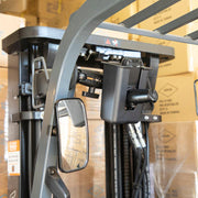 Close-up of a forklift’s overhead guard with the Magnetic Mount and Barcode Scanner Holster Holder, mounted camera system, rearview mirror, and hydraulic hoses—cardboard boxes stacked in the background.