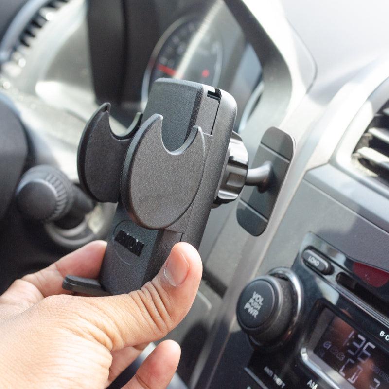 A hand holds the Mega Grip™ Car Phone Holder with Adhesive Mount for iPhone, Galaxy, and Note, ready to attach it to a car’s air vent; dashboard and controls in view—ideal for hands-free use while driving.