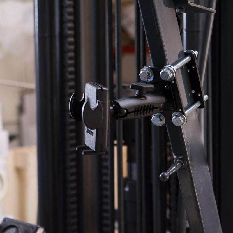 Close-up of the Mega Grip™ Forklift Front Guard Mount securely fastened to a metal frame with bolts and screws, set against blurred industrial equipment in a warehouse.